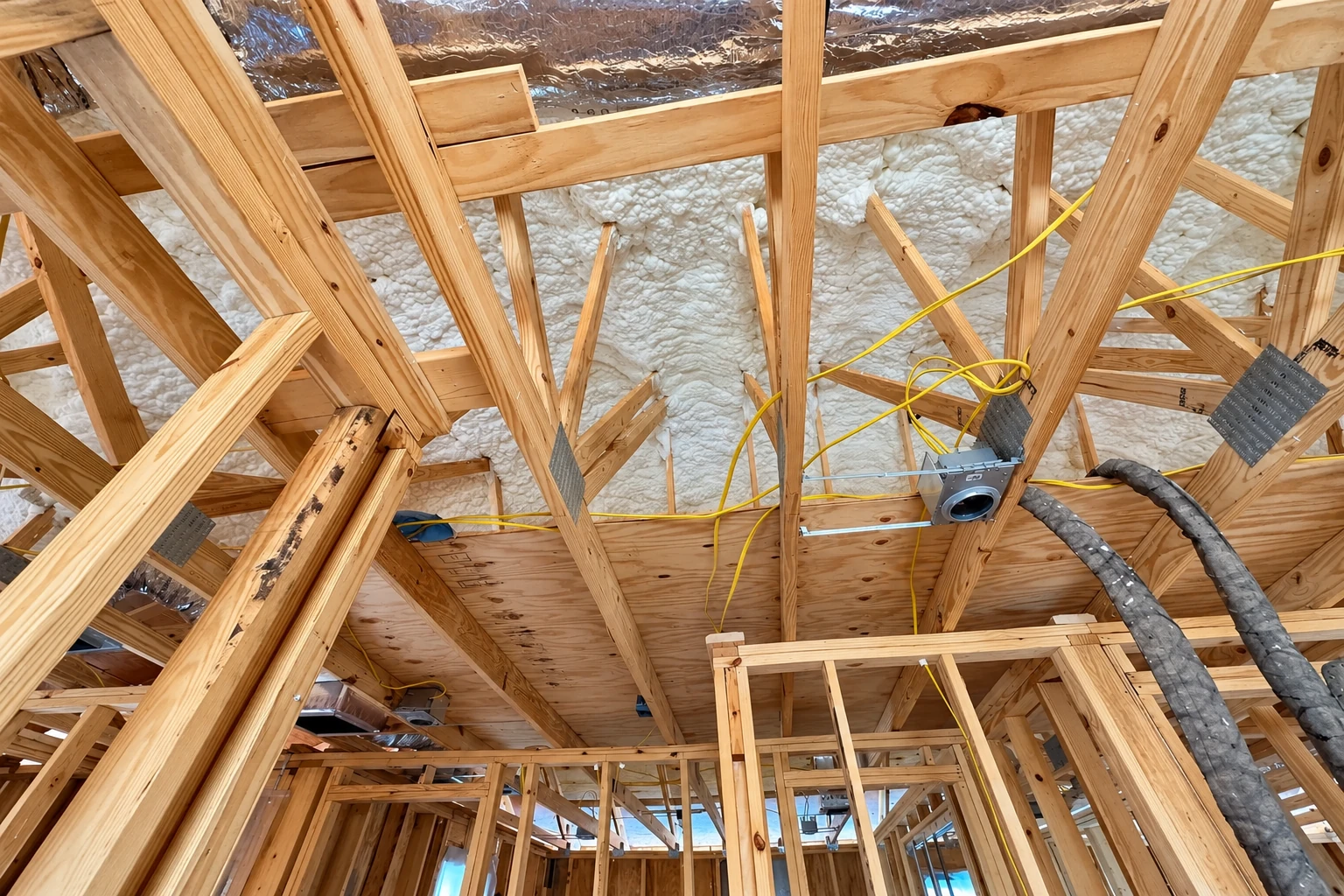 Open roof framing with white spray foam insulation between rafters, yellow wiring, and flexible ducts.