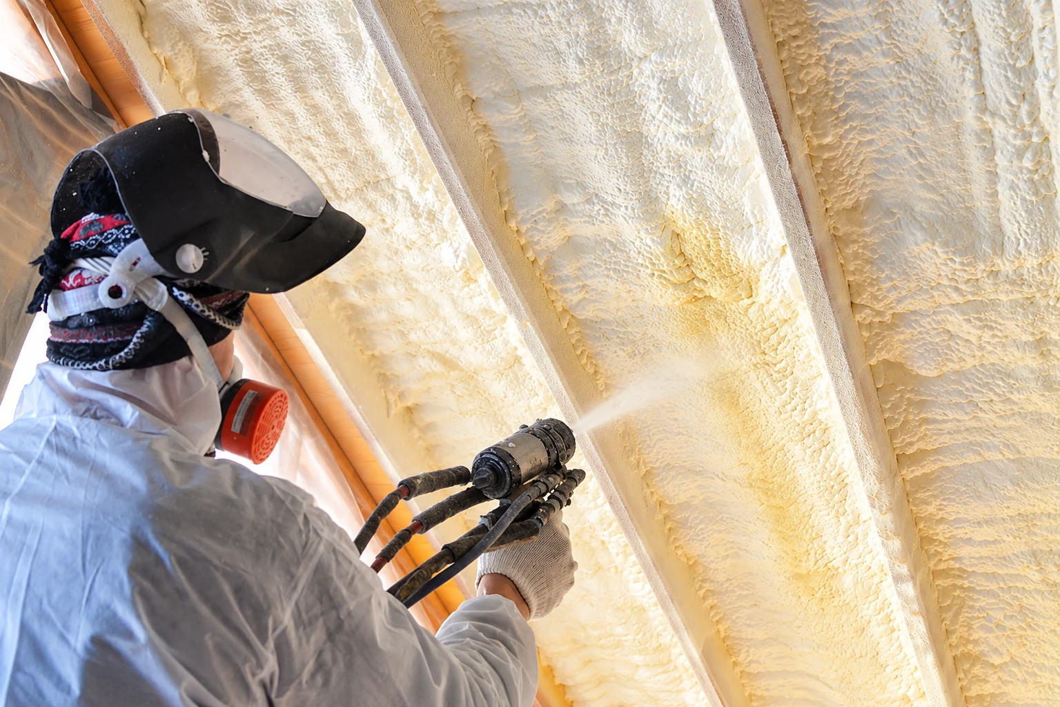 Worker wearing a respirator applies spray foam insulation between wooden ceiling rafters.