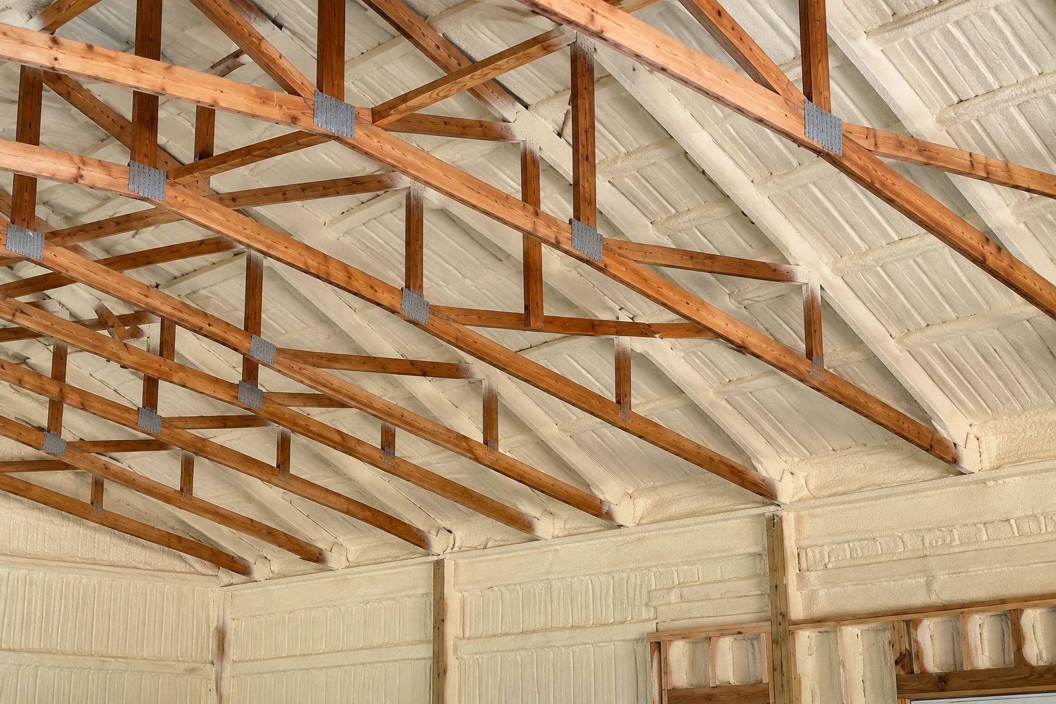 Attic rafters cross beneath a roof coated in pale spray foam insulation.