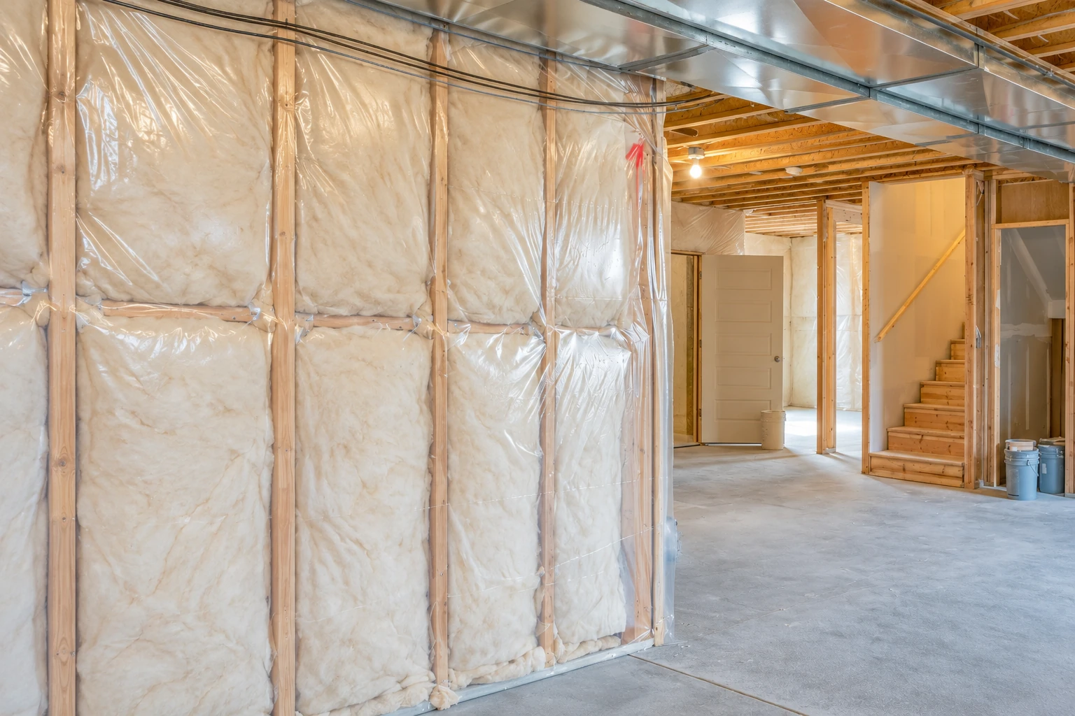 Fiberglass insulation behind clear plastic covers basement wall studs beneath exposed ceiling joists and metal ductwork.