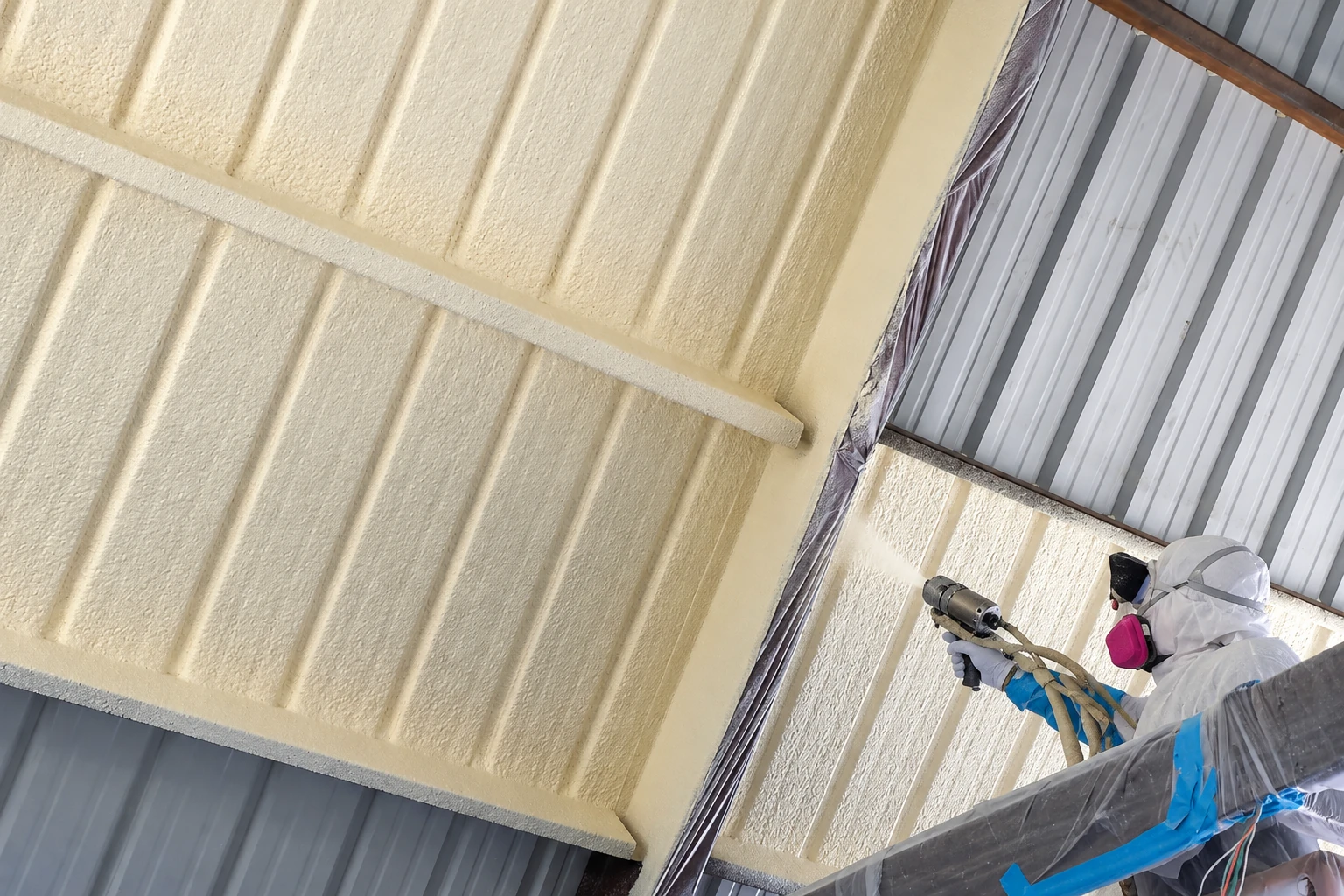 Worker in protective gear sprays foam insulation onto the underside of a corrugated metal commercial roof.