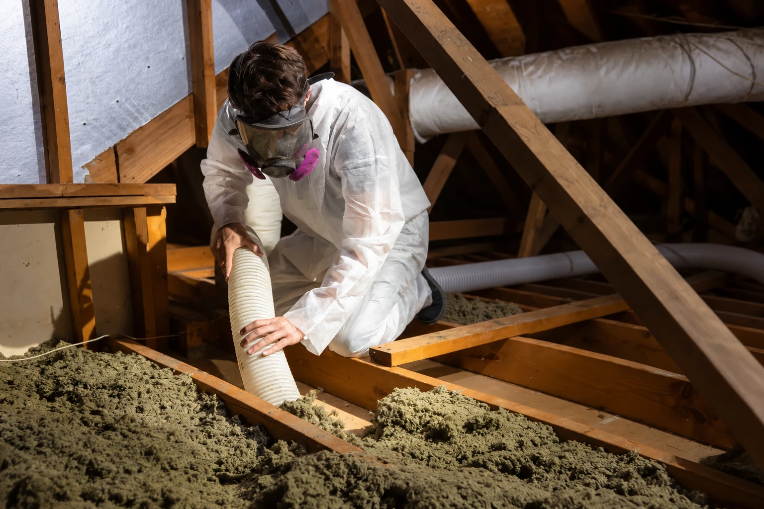 Worker wearing protective coveralls and a respirator uses a hose to remove loose attic insulation.