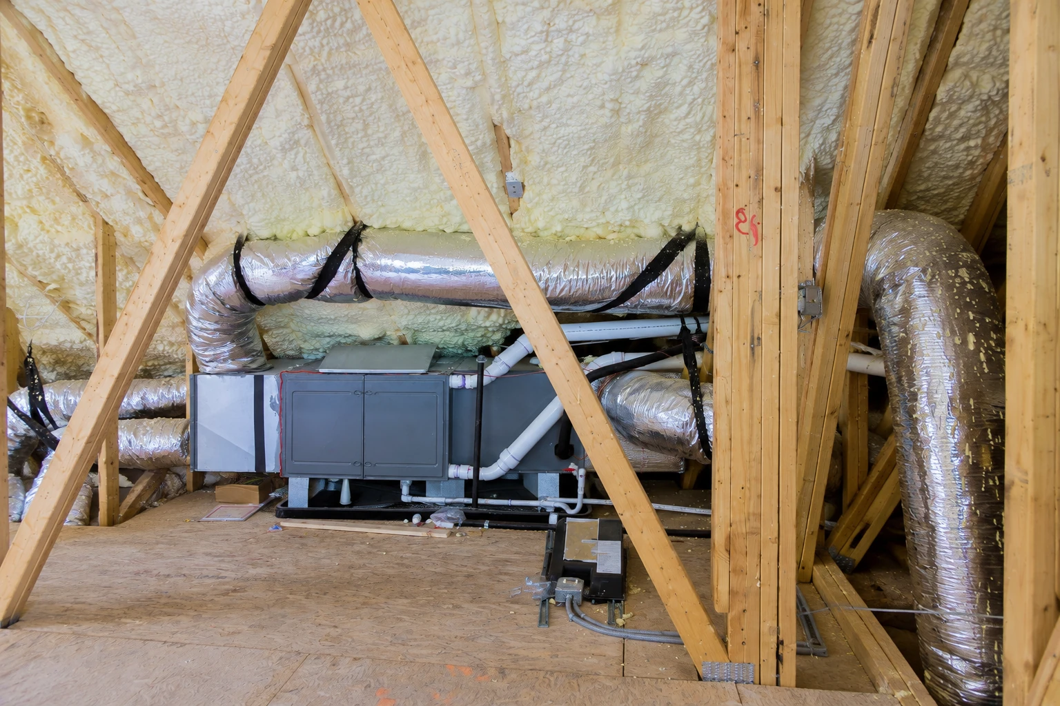 Open-cell foam insulation coats the rafters of an attic containing a central HVAC unit and foil-wrapped ducts.