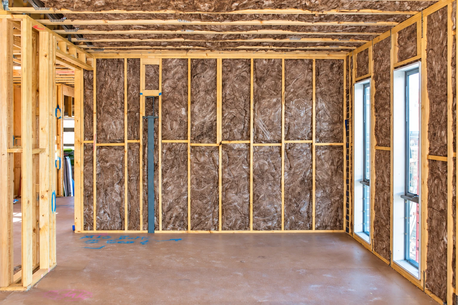 Wall insulation fills the exposed wooden framing in an unfinished room with tall windows and bare flooring.