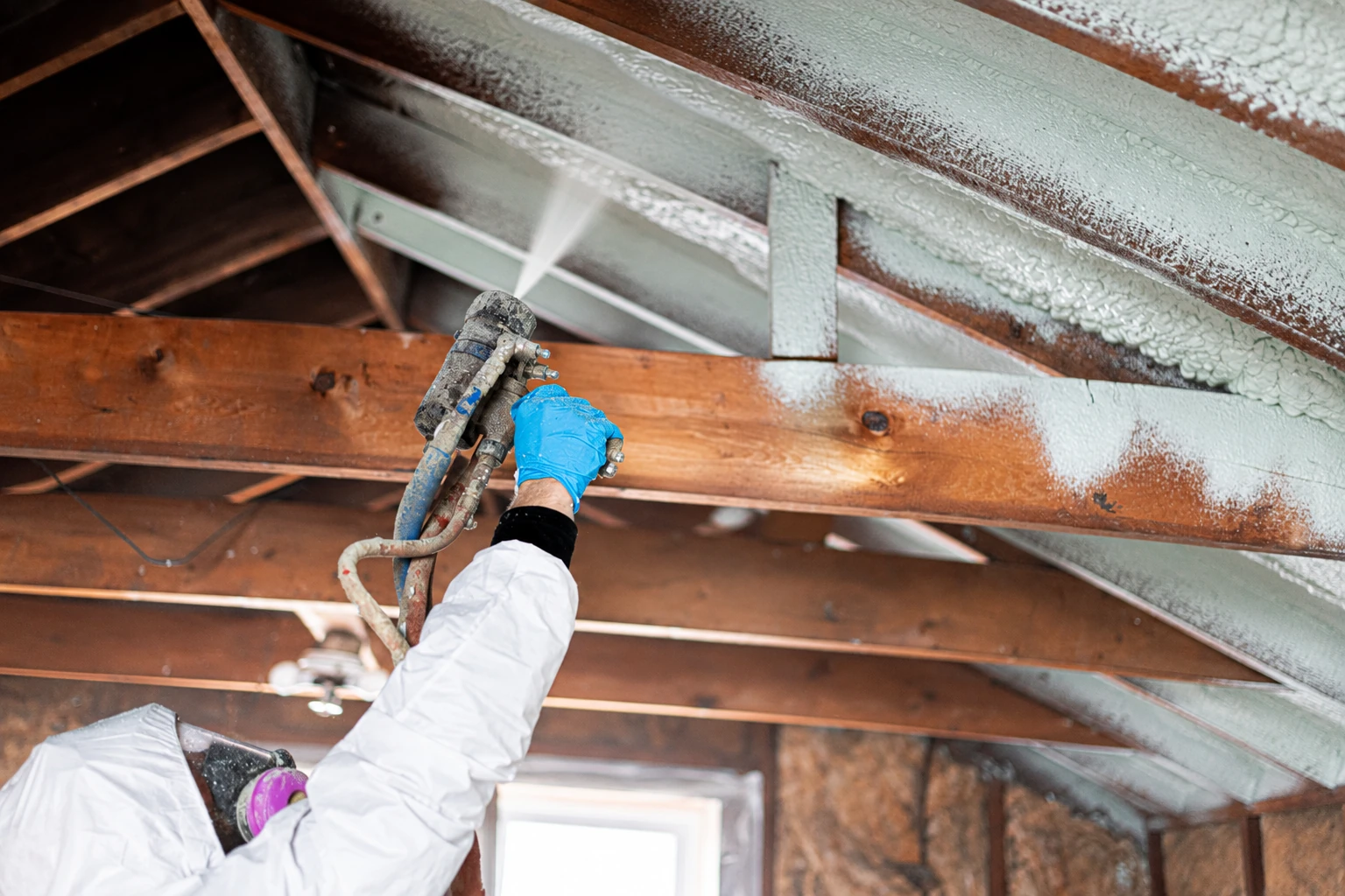 A worker in protective clothing sprays foam insulation onto the underside of a roof between exposed wooden rafters.
